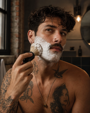 A shirtless man with tattoos applies shaving lather with a brush in a warm, rustic bathroom setting.