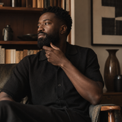 A man with a beard wearing a black short-sleeve button-up shirt sits in a wooden armchair in a warm, book-filled room. He is looking off-camera with his hand resting on his chin.