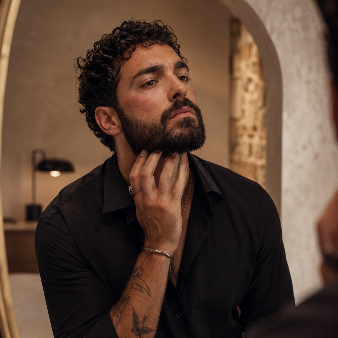 A man with curly dark hair and a beard looks into a mirror while wearing a black button-up shirt. Starting his beard care routine