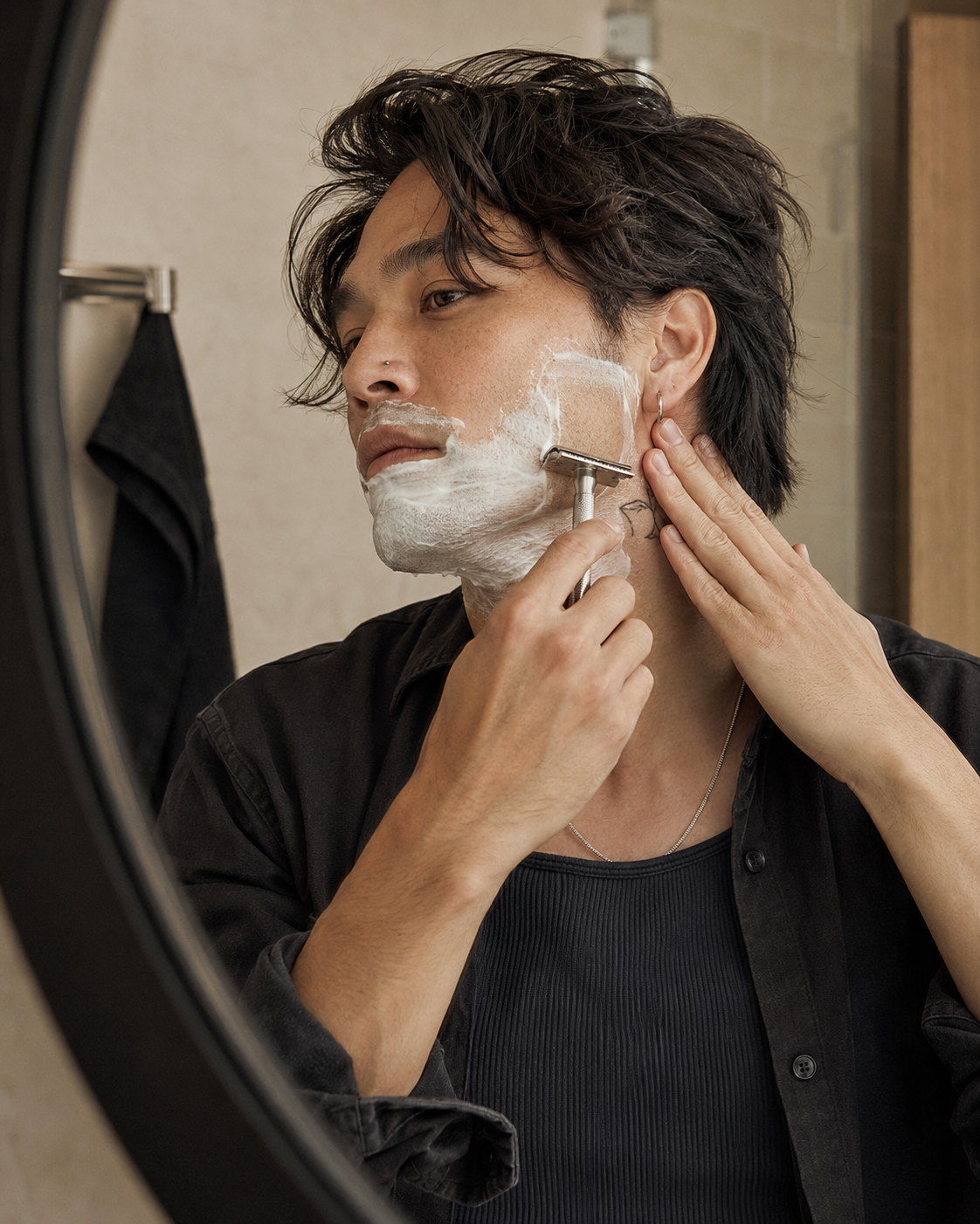 A man with shaving foam on his face uses a silver razor while looking into a bathroom mirror. He is wearing a black button-up shirt over a dark ribbed undershirt.