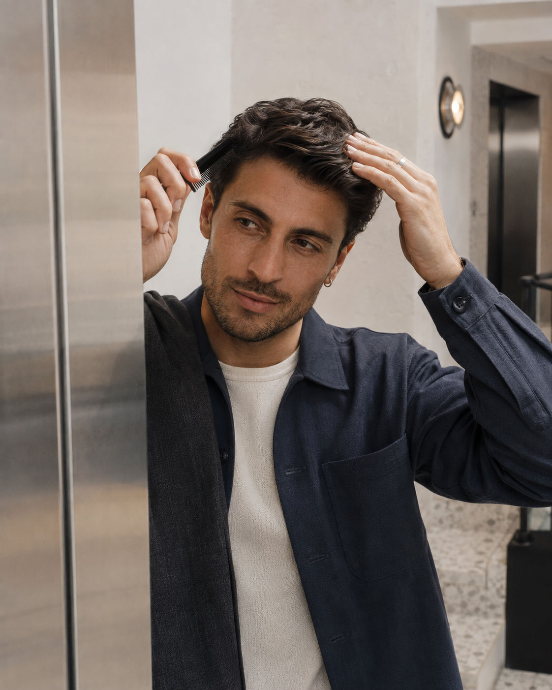 A man in a navy blue overshirt and white t-shirt combing his hair in front of a modern bathroom background.