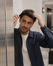 A man in a navy blue overshirt and white t-shirt combing his hair in front of a modern bathroom background.