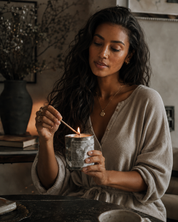A woman with long dark hair lights a candle in a textured beige robe in a cozy, dimly lit room.