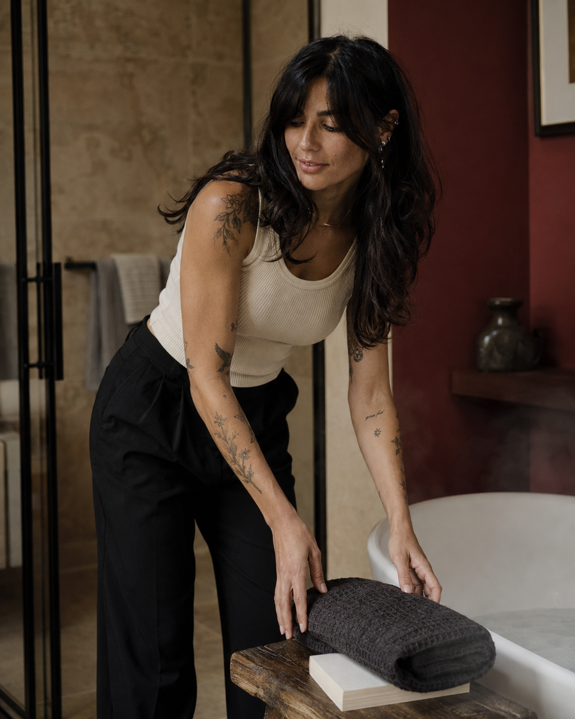 A woman with tattoos leans over a wooden table next to a bathtub, holding a dark charcoal gray textured towel. She is wearing a cream tank top and black trousers in a modern bathroom setting with warm lighting.