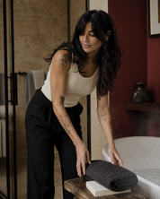 A woman with tattoos leans over a wooden table next to a bathtub, holding a dark charcoal gray textured towel. She is wearing a cream tank top and black trousers in a modern bathroom setting with warm lighting.