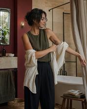 A woman in a bathroom setting wearing an olive green sleeveless top and dark wide-leg pants, while pulling a cream linen shirt off her arms. The bathroom features a terracotta red wall, a stone tub, and a black-framed shower enclosure.