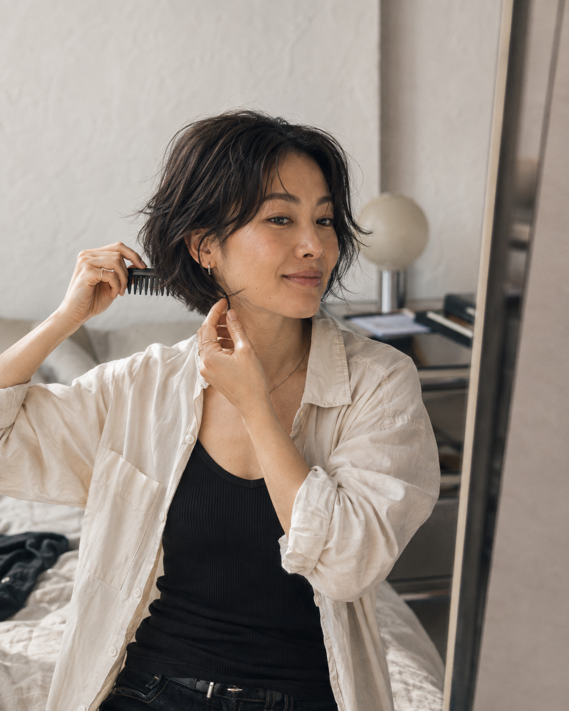 A woman with short dark hair is shown in a bedroom setting, looking into a mirror while combing her hair. She is wearing a black ribbed tank top and an off-white linen button-up shirt.