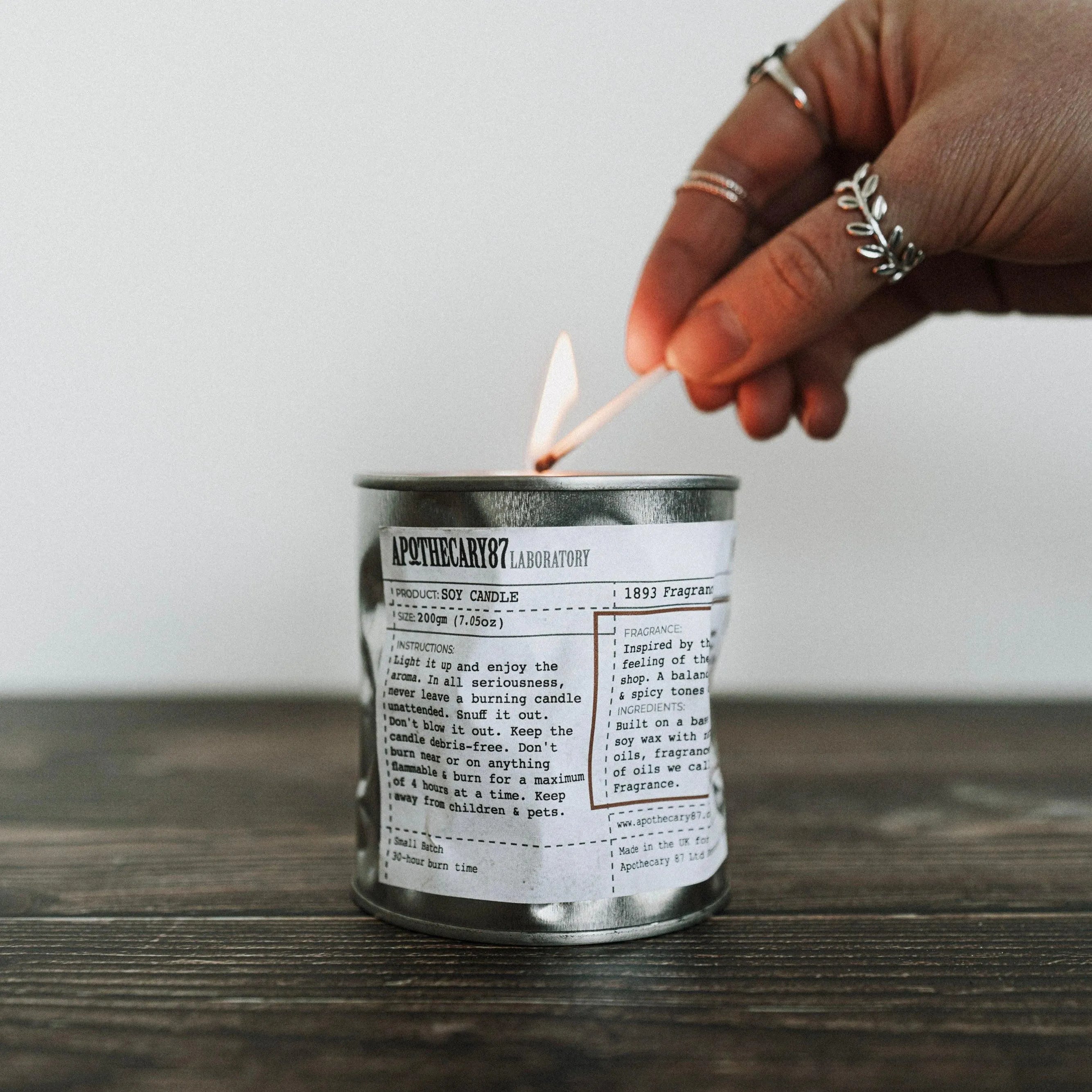 Apothecary 87 Soy Candle distressed tin, being lit with a match. 30+ hours of burn time