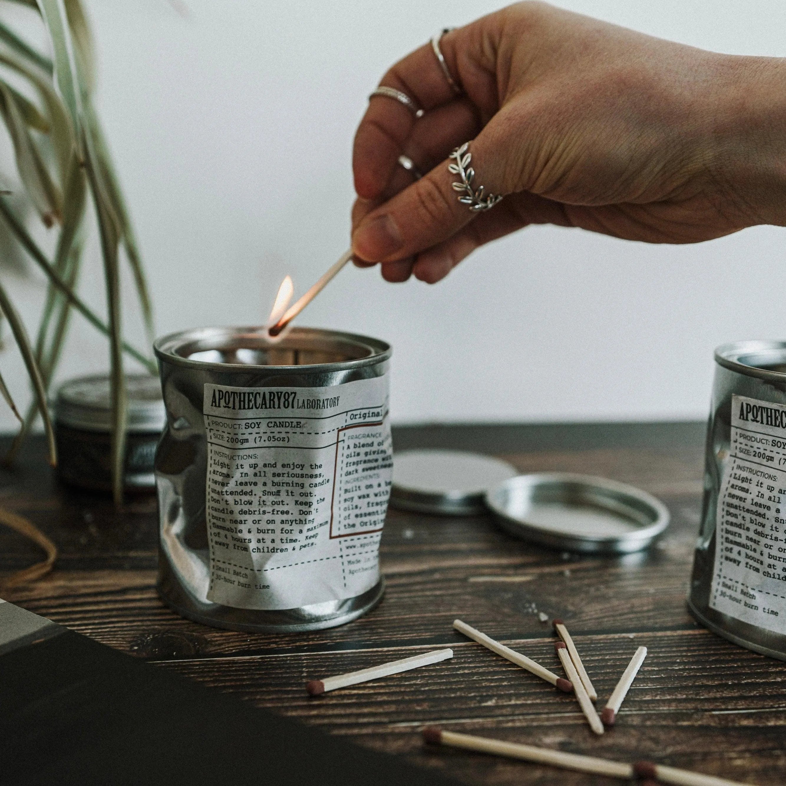 Apothecary 87 Soy Candle crushed tin, being lit with a match. 30+ hours of burn time